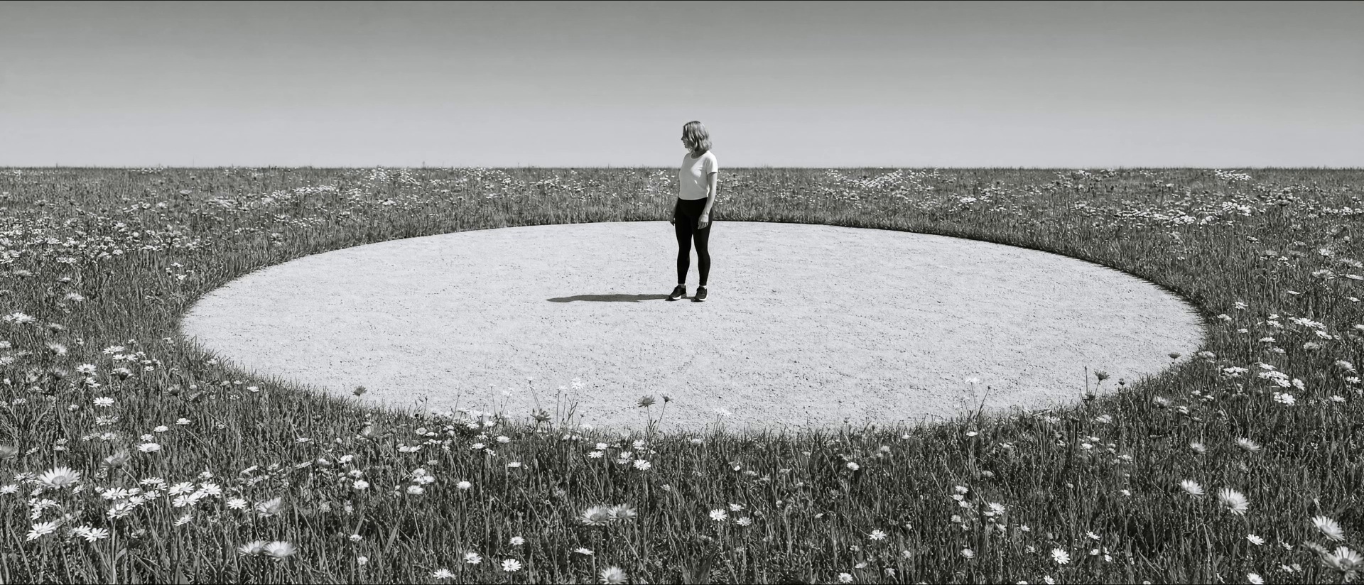 A film still: a lone figure stands in a pale circle cleared in a vast wildflower meadow under an empty sky, in high-contrast black and white.